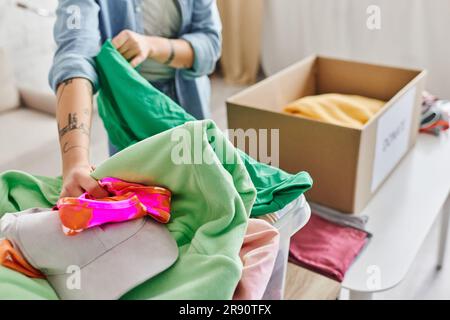 Soziale Initiativen, Teilblick auf junge und tätowierte Frauen, die lässige Kleidung in der Nähe einer Kartonschachtel sortieren, um für eine Sache zu spenden, nachhaltiges Leben Stockfoto