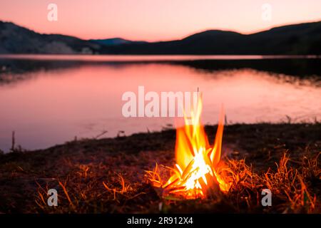 Lagerfeuer. Kleines Lagerfeuer mit sanften Flammen neben einem See während eines glühenden Sonnenuntergangs. San Juan Nacht am Strand. Leute, die über die Lagerfeuer springen. Stockfoto