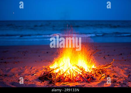 Lagerfeuer. Kleines Lagerfeuer mit sanften Flammen neben einem See während eines glühenden Sonnenuntergangs. San Juan Nacht am Strand. Leute, die über die Lagerfeuer springen. Stockfoto