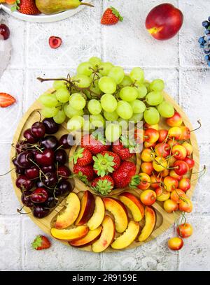 Reichhaltiger Obstteller gefüllt mit Früchten und Beeren. Auswahl an schwarzer Kirsche, grüner Traube, gelber Kirsche, Pfirsichen und Erdbeere auf Holzplatte auf dem Stockfoto