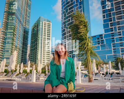 Junge Frau im grünen Anzug geht durch die Straßen von Dubai, Dubai Marina District. Reisekonzept der Vereinigten Arabischen Emirate. Die Vorstellung von erfolgreichem Auswanderer, Umzug in ein anderes Land, Arbeitsvisum Stockfoto