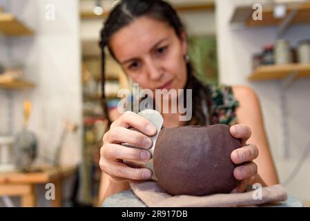 Nahaufnahme der Hände einer Frau, die rohe Keramik und Lehm bearbeitet, um Topf oder Schüssel zu machen. Keramikwerkstatt mit natürlichem Licht zum Schaben, Glätten, Formen und Glätten Stockfoto
