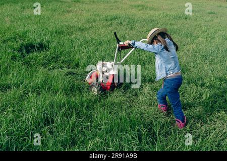 Ein kleines, süßes Mädchen, das einen Rasenmäher auf ein riesiges Grasfeld schiebt Stockfoto