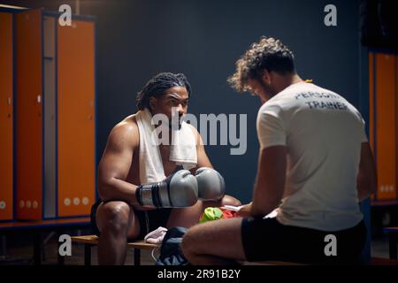 Breite Horizontalaufnahme eines starken, muskulösen Boxspielers nach dem Training in Schweiß, der mit seinem Trainer auf der Bank sitzt und spricht. Stockfoto