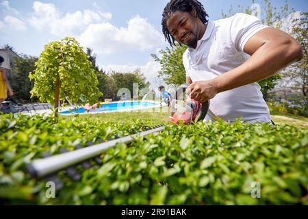 Der Mensch schwingt furchtlos mit einem Trimmer, während er eine lebende Hecke mit größter Sorgfalt formt. Mit zielgerichteter Entschlossenheit navigiert er fachkundig durch das dichte Laub, Stockfoto