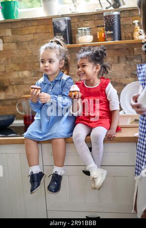 Zwei junge Mädchen werden in einem wunderbaren Moment gefangen, während sie auf der Küchentheke sitzen und köstliche Muffins mit Muffins genießen. Mit einem Lächeln der reinen Freude Stockfoto