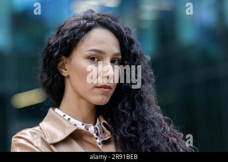 Porträt einer erwachsenen Geschäftsfrau außerhalb des Bürogebäudes, einer lateinamerikanischen Frau mit lockigem Haar, die in die Kamera schaut, einer Chefin in Geschäftskleidung. Stockfoto