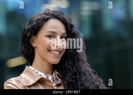 Nahaufnahme eines jungen, wunderschönen hispanischen Geschäftsmanns, der mit lockigem Haar vor dem Bürogebäude lächelt und in die Kamera schaut. Der Finanzier ist mit den erfolgreichen Ergebnissen zufrieden. Stockfoto