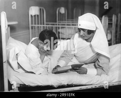 New York, New York: 5. Januar 1926 Mädchen der New Yorker Gesellschaft verbringen einen Tag pro Woche als ehrenamtliche Krankenschwestern im Dienst der Menschheit. Miss Betty Nixon sieht einem Jungen im Tonsil Hospital in Manhattan vorlesen. Stockfoto