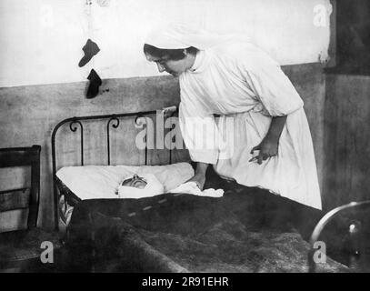 Frankreich: Am 21. November 1914 Neigt Eine französische Krankenschwester zu einem schwer verwundeten belgischen Soldaten, der seiner Frau einen Brief über die Babyschuhe geschrieben hatte, die er für seinen dreijährigen Sohn gekauft hatte, mit seinem Einkommen aus seiner Tätigkeit als Pfadfinder in König Alberts Armee. Die Schuhe hängen an der Wand über seinem Kopf. Stockfoto