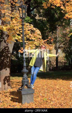 Das Foto wurde in der Stadt Odessa gemacht. Auf dem Bild steht ein freundliches Mädchen in einem gelben Regenmantel im Herbst auf dem Fuß eines Straßenlaterenpfeilers Stockfoto