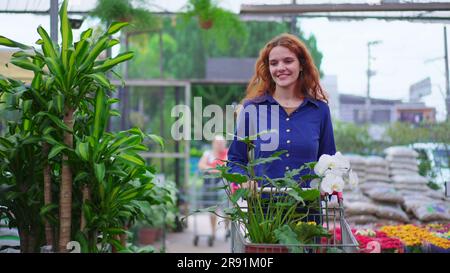 Freudige Frau, die Pflanzen und Blumen mit einem Wagen in einem Blumenladen auswählt. Weibliche Kunden, die in einem Geschäft vor Ort einkaufen Stockfoto