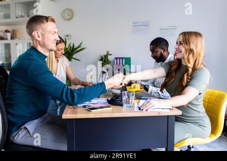 Lächelnde Geschäftskollegen, die sich in einer Bürobesprechung am Schreibtisch die Hand schütteln Stockfoto