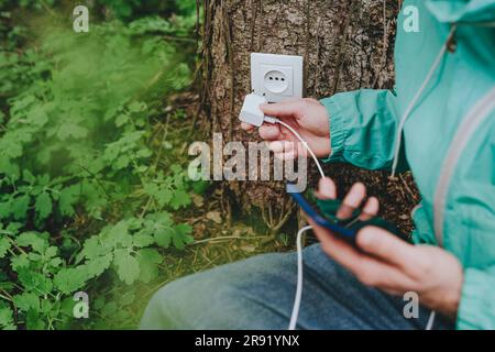 Mann, der ein Mobiltelefon-Ladegerät in die Steckdose am Baumstamm einsteckt Stockfoto