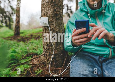 Mann, der ein Smartphone benutzt, das mit einer Steckdose am Baumstamm verbunden ist Stockfoto