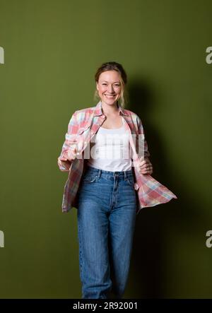 Lächelnde junge Frau in einem karierten Hemd, die vor einer grünen Wand steht Stockfoto