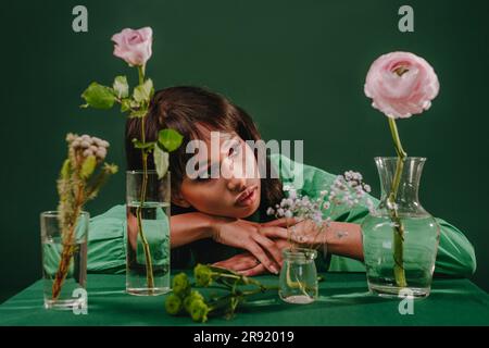 Rücksichtsvolle Frau, die sich auf den Tisch lehnt und Blumen vor dem grünen Hintergrund trägt Stockfoto