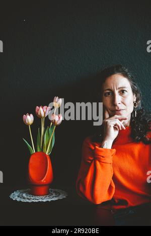 Frau mit der Hand am Kinn, die sich auf den Tisch lehnt, mit roter Blumenvase vor der schwarzen Wand Stockfoto