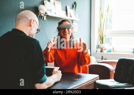 Glückliche Frau, die ein VR-Headset trägt und von einem Mann zu Hause gesessen wird Stockfoto