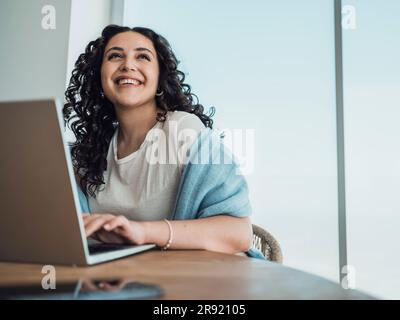 Fröhliche Frau, die aufblickte und am Tisch ein Notebook benutzte Stockfoto