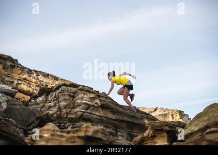 Junge Frau, die unter dem Himmel auf felsige Hügel klettert Stockfoto