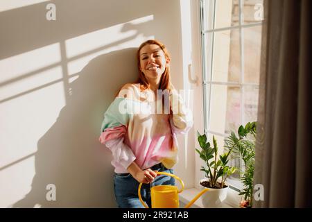 Lächelnde rothaarige Frau mit Gießkanne bei Pflanzen auf Fensterbank Stockfoto