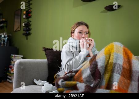 Eine kranke junge Frau, die sich auf der Couch entspannt, mit Tüchern die Nase bläst Stockfoto