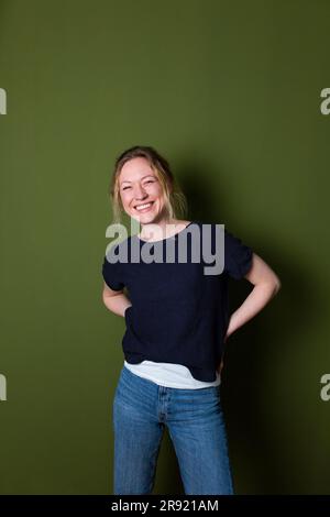 Lächelnde junge Frau in blauem T-Shirt, die mit den Händen hinter dem Rücken an der grünen Wand steht Stockfoto