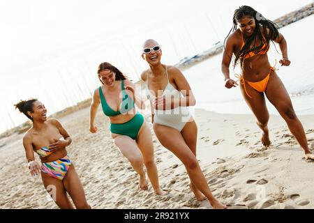 Fröhliche Freunde, die Bikinis tragen und am Strand auf Sand laufen Stockfoto