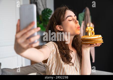 Junge Frau bläst Kerzen auf Geburtstagskuchen und macht Selfie zu Hause Stockfoto