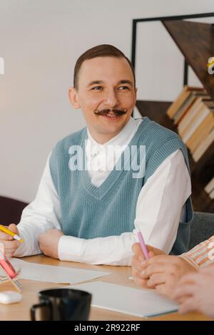 Geschäftsmann mit Schnurrbart, sitzt am Tisch und hört Kollegen zu Stockfoto