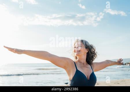 Lächelnde reife Frau, die Yoga mit ausgestreckten Armen am Strand praktiziert Stockfoto