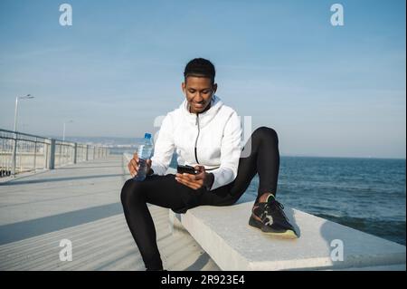 Lächelnder Sportler mit Smartphone, der auf einer Bank in der Nähe des Meeres sitzt Stockfoto