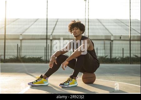 Selbstbewusster Sportler sitzt auf dem Basketball auf dem Sportplatz Stockfoto