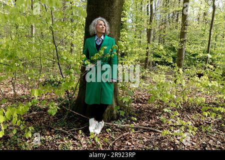 Seniorin lehnt sich im Wald an einen Baum und trägt einen grünen Mantel Stockfoto