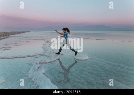 Junge Frau, die bei Sonnenuntergang auf Salzformationen im toten Meer springt Stockfoto