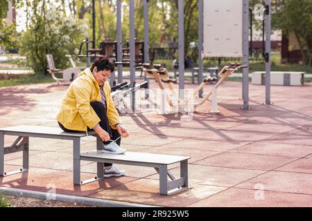 Seniorin, die Schnürsenkel bindet, sitzt auf einer Bank im Park Stockfoto