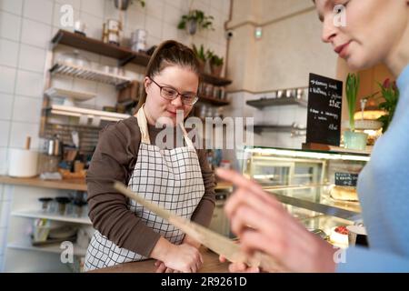 Lächelnder Cafebesitzer mit Down-Syndrom, der Gästen hilft, Speisen von der Speisekarte auszuwählen Stockfoto