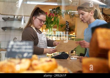 Der Cafebesitzer mit Down-Syndrom hilft dem Gast, Speisen von der Speisekarte im Café zu bestellen Stockfoto