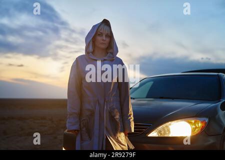 Eine blonde Frau in einem Regenmantel, die neben einem beleuchteten Autoscheinwerfer steht Stockfoto