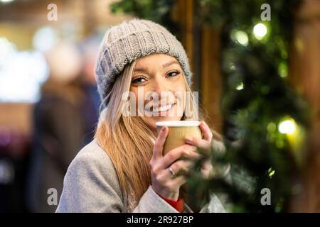 Lächelnde junge Frau, die eine Kaffeetasse auf dem Weihnachtsmarkt hält Stockfoto