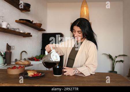 Junge Frau, die frisch gebrühten Kaffee in eine Tasse am Küchentisch einschenkt Stockfoto