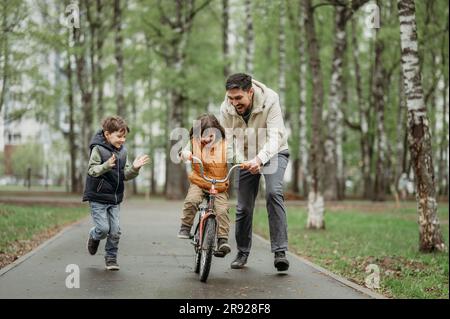Ein jubelnder Bruder lernt Fahrrad mit Vater im Park zu fahren Stockfoto
