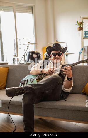 Lächelnder Gitarrist mit Hut und Gitarre zu Hause Stockfoto