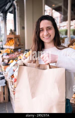 Ein glücklicher Lebensmittelhändler steht mit einer Papiertüte am Gemüsestand Stockfoto