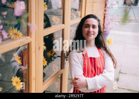 Lächelnder Blumenhändler mit Schürze, der mit verschränkten Armen steht Stockfoto
