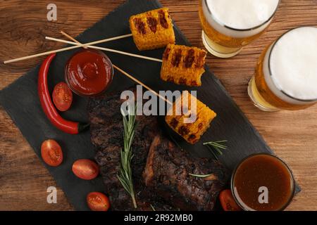 Gläser Bier, leckere gegrillte Rippchen, Mais und Soßen auf Holztisch, flach liegend Stockfoto
