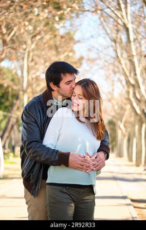 Ein Mann küsst seine glückliche schwangere Frau, ein Paar umarmt und berührt ihren Bauch in einem Park. Stockfoto