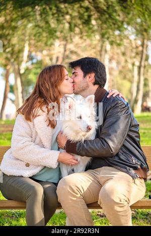 Ein junges weißes Paar küsst sich mit seinem Hund, der auf einer Bank in einem Park sitzt. Familie. Stockfoto