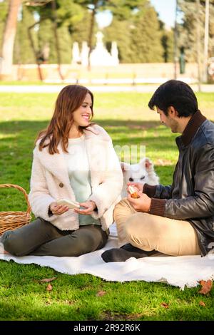 Ein junges Paar sieht sich an und isst ein Sandwich auf einem Picknick in einem Park mit seinem Hund. Stockfoto